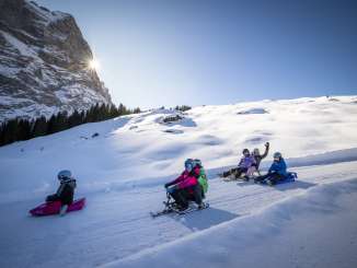 Mehrere Kinder fahren mit unterschiedlichen Schlitten gemeinsam die Piste hinunter. Die Szene zeigt verschiedene Fahrpositionen und Abstände innerhalb der Gruppe. Im Hintergrund ragt die Sonne über den Mättenberg.