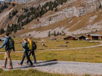 Three hikers are walking along a wide gravel path through an open Alpine valley. Several traditional wooden huts stand on the right, while rocky mountain slopes frame the valley. The light has a clear, late-autumnal quality.