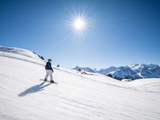 HaslibergEine Snowboardfahrerin gleitet über die Piste auf dem Hasliberg und zieht ihre Spur durch den Schnee. Die Sonne steht hoch am Himmel und wirft lange Schatten auf die Oberfläche. Im Hintergrund erstreckt sich die winterliche Berglandschaft.