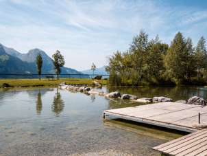 A wooden pier juts out into the clear Hasliberg swimming lake, whose shores are lined with rocks and trees. In the background, the view opens up to the surrounding mountains, while a few trees and a bench stand on the meadow. The calm surface of the water reflects the summer landscape.
