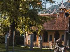 La statue en bronze de Sherlock Holmes est assise sur une pierre dans un parc, près d'un cours d'eau. À l'arrière-plan, on aperçoit le bâtiment du musée, des panneaux d'information, ainsi que des arbres aux feuilles d'automne et un paysage de montagne.