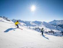 Two skiers are skiing down a wide, snow-covered slope on the Hasliberg. The sun is high in the sky, casting long shadows on the snow. In the background, other mountain ranges and snow-capped peaks are visible.