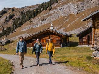 Drei Wandernde gehen einen breiten Schotterweg entlang, flankiert von traditionellen Holzhütten. Die umliegenden Felswände bilden eine markante Kulisse. Die Lichtstimmung ist klar und herbstlich.