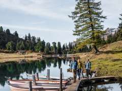 Three people are walking across a narrow wooden walkway with several rowboats moored alongside it. Lake Engstlen clearly reflects the surrounding trees and the sky. The surroundings have an autumnal and peaceful feel.