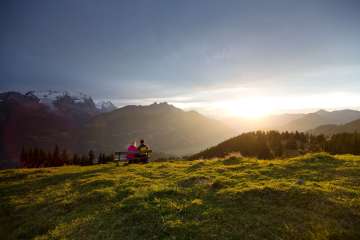 Zwei Personen sitzen auf einer Bank oberhalb der Mägisalp und blicken in die Abendsonne. Das warme Licht taucht die Wiesen und Berglandschaft des Haslibergs in goldene Farben, während die Gipfel der Berner Alpen die Kulisse bilden.