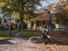 Une statue en bronze de Sherlock Holmes est assise sur une pierre dans un parc. À l'arrière-plan, on aperçoit des panneaux d'information, des arbres aux feuilles d'automne et les bâtiments du site du musée.