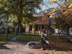 Die Bronzestatue von Sherlock Holmes sitzt auf einem Stein in einer Parkanlage neben einem Wasserlauf. Im Hintergrund stehen das Museumsgebäude, Informationstafeln sowie Bäume mit Herbstlaub und Berglandschaft.