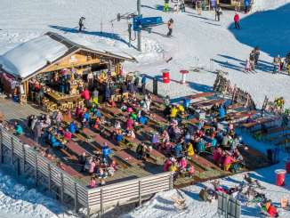 Die Kuhstallbar auf der Mägisalp ist von zahlreichen Gästen besucht, die auf der Sonnenterrasse sitzen. Rund um die Bar verlaufen Skipisten, auf denen Wintersportler unterwegs sind. Die Anlage liegt mitten im Skigebiet des Haslibergs.