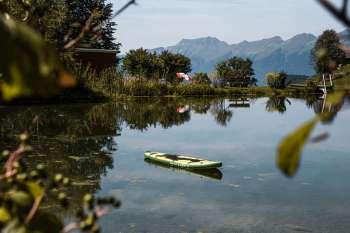 Une planche de stand up paddle verte flotte tranquillement sur les eaux limpides du lac de baignade de Hasliberg. Sur la rive, on aperçoit des chaises longues et un parasol coloré, tandis qu’en arrière-plan, les montagnes dominent le paysage. Les arbres et les buissons se reflètent dans l’eau et encadrent ce tableau idyllique.