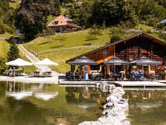 On the shore of Hasliberg Lake stands a cozy wooden house with a patio and umbrellas. Visitors enjoy the warm weather while the surrounding landscape is reflected in the clear water. Green hillsides dotted with trees and scattered houses provide an idyllic backdrop.