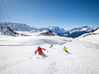 Three skiers are skiing down a wide slope on the Hasliberg. Snow is kicked up as they turn. In the background, the Wetterhorn, the Engelhörner, the Eiger, and other mountain peaks are visible.