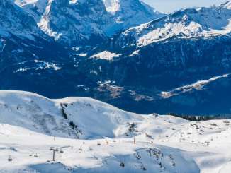 The snow-covered landscape of the Hasliberg stretches across gentle hills and slopes. At the bottom center of the picture lies the Mägisalp with its buildings, while the Eiger, the Wetterhorn, the Grosse Scheidegg, and other mountain peaks are visible in the background.