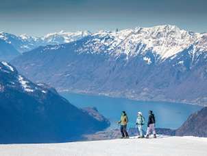 Zwei Skifahrer und eine Snowboarderin stehen auf dem Hasliberg und blicken über die Berglandschaft. Im Hintergrund ist der Brienzersee zwischen den umliegenden Bergketten gut sichtbar.