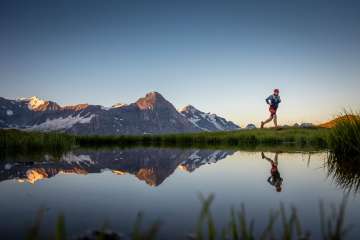 A runner is running along the mountain lake. The mountain panorama, including the Eiger, is reflected in the mountain lake in the morning sun.