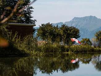 A green stand-up paddleboard floats peacefully on the clear waters of Lake Hasliberg. Deck chairs and a colorful beach umbrella are set up on the shore, while the mountains dominate the scenery in the background. Trees and shrubs are reflected in the water, framing the idyllic scene.