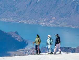 Deux skieurs et une snowboardeuse se tiennent sur le Hasliberg et contemplent le paysage montagneux. À l'arrière-plan, on aperçoit clairement le lac de Brienz entre les chaînes de montagnes environnantes.