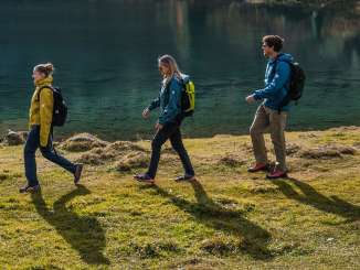 The group walks along the lakeshore; the water shimmers in a vivid turquoise. The autumn foliage and the shade of the trees create an atmospheric scene.