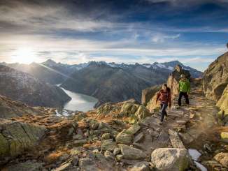 Deux randonneurs en route dans l'univers du Grimsel.  Ils marchent  devant une vue imprenable sur le lac de barrage du Grimsel, dans l'univers du Grimsel. Sur le sentier de randonnée entre le col du Grimsel et le Nägelisgrätli.  