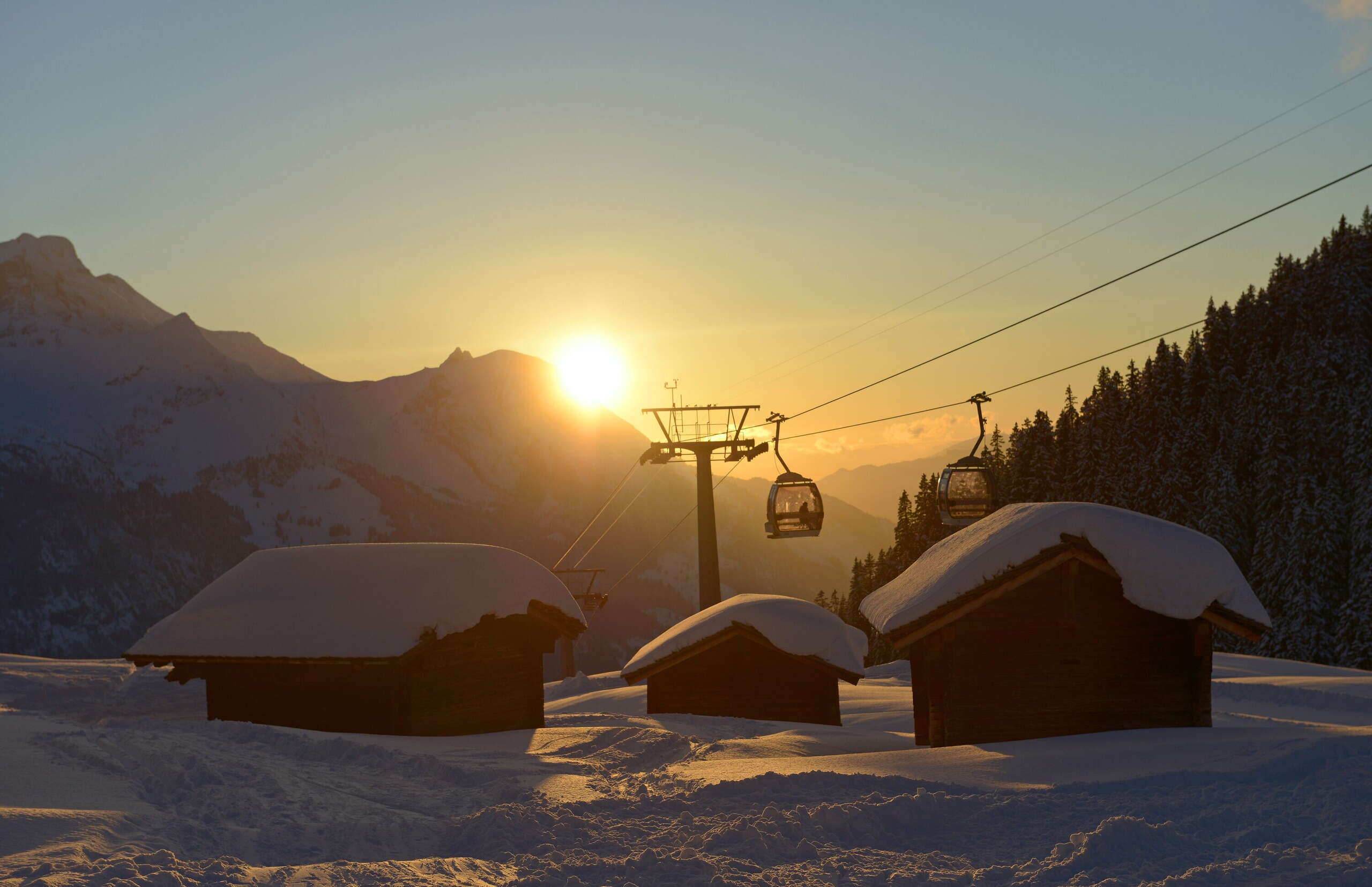 Deux télécabines des remontées mécaniques de Meiringen-Hasliberg au coucher du soleil, avec au premier plan trois cabanes enneigées. Prise de vue à la station intermédiaire de Bidmi.