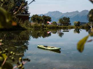 Ein grünes Stand-Up-Paddle treibt ruhig auf dem klaren Badesee Hasliberg. Am Ufer stehen Liegestühle und ein bunter Sonnenschirm, während im Hintergrund die Berge die Szenerie dominieren. Bäume und Sträucher spiegeln sich im Wasser und rahmen das idyllische Bild ein.