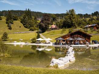 On the shore of Hasliberg Lake stands a cozy wooden house with a terrace and umbrellas. Visitors enjoy the warm weather while the surroundings are reflected in the clear water. Green hillsides dotted with trees and scattered houses form the idyllic backdrop.