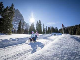 Zwei Kinder fahren gemeinsam auf einem Schlitten die Schlittelpiste von der Grossen Scheidegg zur Schwarzwaldalp hinunter. Die tief stehende Sonne sorgt für starkes Gegenlicht und lange Schatten auf dem Schnee. Im Hintergrund ist das Wetterhorn sichtbar, während weitere Kinder mit Abstand folgen.