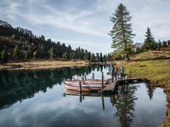 Trois personnes marchent sur une étroite passerelle en bois, à laquelle sont amarrées plusieurs barques. Le lac d'Engstlen reflète clairement les arbres environnants et le ciel. Le cadre dégage une atmosphère automnale et paisible.