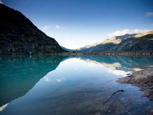 Le lac de Gelmer est niché au cœur du paysage de haute montagne du massif du Grimsel. La surface calme de l'eau reflète les flancs des montagnes environnantes et le ciel limpide. Au premier plan, on aperçoit les rochers des rives, tandis qu'au fond, le barrage délimite le lac.