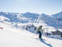 Two skiers are skiing down a wide, snow-covered slope on the Hasliberg. The sun is high in the sky and casts long shadows on the snow. In the background, other mountain ranges and snow-capped peaks are visible.