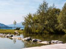 Une jetée en bois s'avance dans le lac de baignade aux eaux claires de Hasliberg, dont les rives sont bordées de pierres et d'arbres. À l'arrière-plan, la vue s'ouvre sur les montagnes environnantes, tandis que la prairie est parsemée d'arbres isolés et d'un banc. La surface calme de l'eau reflète le paysage estival.