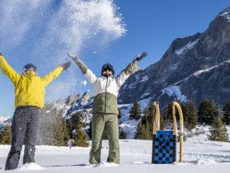 Two people are tossing snow into the air while standing on a snow-covered area. Several sleds are parked in the snow next to them. The Wellhorn rises in the background.
