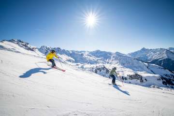 Two skiers are skiing down a wide, snow-covered slope on the Hasliberg. The sun is high in the sky and casts long shadows on the snow. In the background, other mountain ranges and snow-capped peaks are visible.