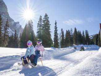 Two children are sledding together down the sledding run from the Grosse Scheidegg to the Schwarzwaldalp. The low-hanging sun creates a strong backlight and long shadows on the snow. The Wetterhorn is visible in the background, while other children follow at a distance.
