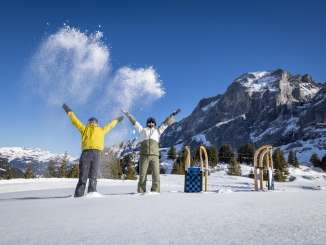 Zwei Personen werfen Schnee in die Luft und stehen dabei auf einer verschneiten Fläche. Neben ihnen stehen mehrere Schlitten im Schnee. Im Hintergrund erhebt sich das Wellhorn.