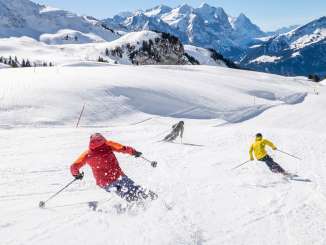 Three skiers are skiing down a wide slope on the Hasliberg. Snow is kicked up as they turn. In the background, the Wetterhorn, the Engelhörner, the Eiger, and other mountain peaks are visible.