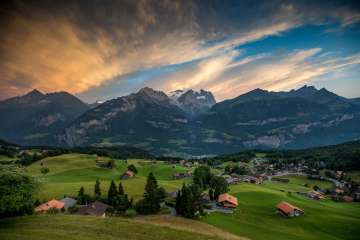 Die Häuser stehen in den grünen Wiesen am Hasliberg. Das Bergpanorama im Hintergrund wird von Wolken überdeckt. Der Sonnenuntergang lässt die Wolken orange leuchten.