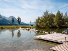 Ein Holzsteg ragt in den klaren Badesee Hasliberg, dessen Ufer von Steinen und Bäumen gesäumt ist. Im Hintergrund öffnet sich der Blick auf die umliegenden Berge, während auf der Wiese einzelne Bäume und eine Bank stehen. Die ruhige Wasseroberfläche spiegelt die sommerliche Landschaft.