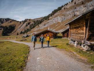 Drei Wandernde gehen einen breiten Schotterweg entlang, flankiert von traditionellen Holzhütten. Die umliegenden Felswände bilden eine markante Kulisse. Die Lichtstimmung ist klar und herbstlich.