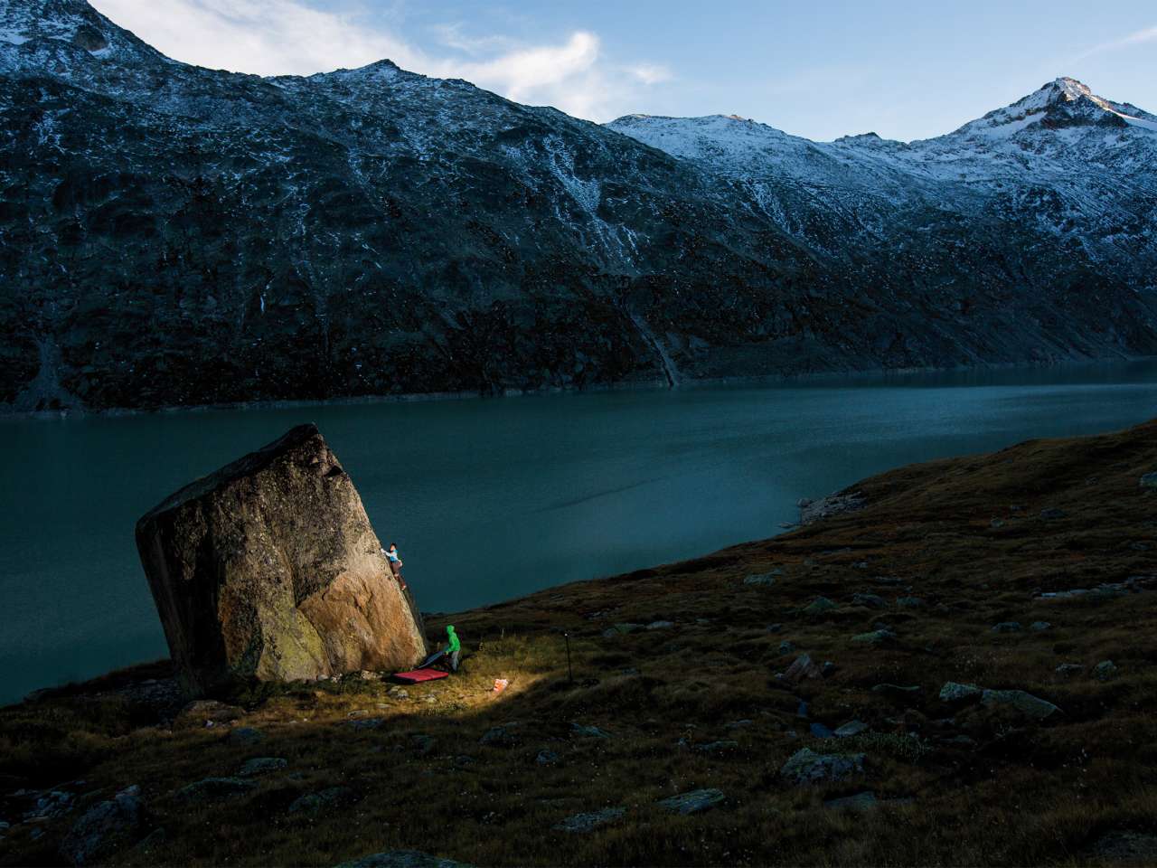 Zwei Kletterer an einem Kletterfelsen in der Dämmerung mit einem See und Bergen im Hintergrund.
