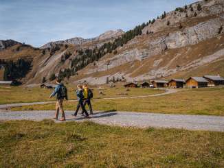 Three hikers are walking along a wide gravel path through an open Alpine valley. Several traditional wooden huts stand on the right, while rocky mountain slopes frame the valley. The light has a clear, late-autumnal quality.