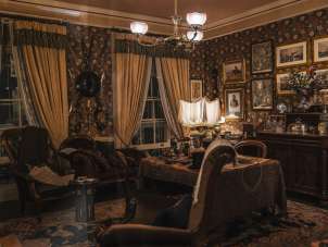 A reconstructed interior at the Sherlock Holmes Museum, featuring a desk, armchairs, a rug, lamps, and display cases. Historical paintings and decorations hang on the walls. The room is designed in the style of a Victorian study.