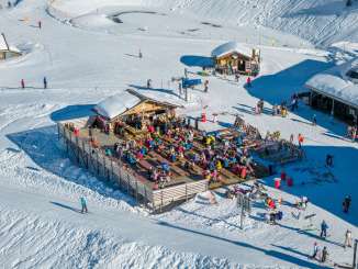 Die Kuhstallbar auf der Mägisalp ist von zahlreichen Gästen besucht, die auf der Sonnenterrasse sitzen. Rund um die Bar verlaufen Skipisten, auf denen Wintersportler unterwegs sind. Die Anlage liegt mitten im Skigebiet des Haslibergs.