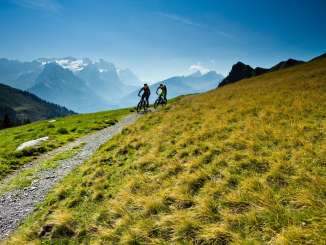 Two mountain bikers are riding along a mountain trail through the alpine landscape of the Hasliberg. The peaks of the Bernese Alps are visible in the background, while sunny mountain meadows dominate the scene.