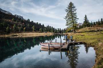 Drei Personen gehen über einen schmalen Holzsteg, an dem mehrere Ruderboote festgemacht sind. Der Engstlensee spiegelt die umliegenden Bäume und den Himmel klar wider. Die Umgebung wirkt herbstlich und ruhig.