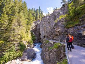 Der Fluss formte die Rosenlaui Gletscherschlucht über Jahrhunderte. Der Weg führt an der Felswand über dem Fluss in die Schlucht hinein. Zwei Wanderer bestaunen den Fluss vor dem Eingang in die Schlucht.