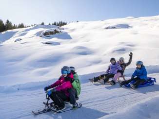 Several children are sledding down the slope together on different sleds. The scene shows various riding positions and distances between the children within the group. In the background, the sun rises over the Mättenberg.