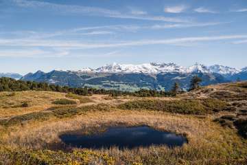 Ein kleiner Bergsee liegt eingebettet in eine karge, von Sträuchern und Gras bewachsene Landschaft. Im Hintergrund sind schneebedeckte Gipfel und blauer Himmel zu sehen.